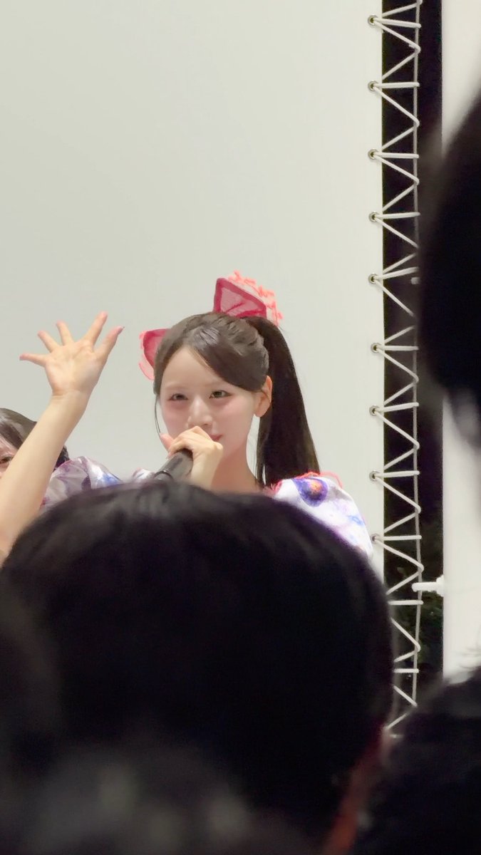A young woman with a pink bow in her ponytail holds a microphone and waves to an audience during what appears to be a live performance.