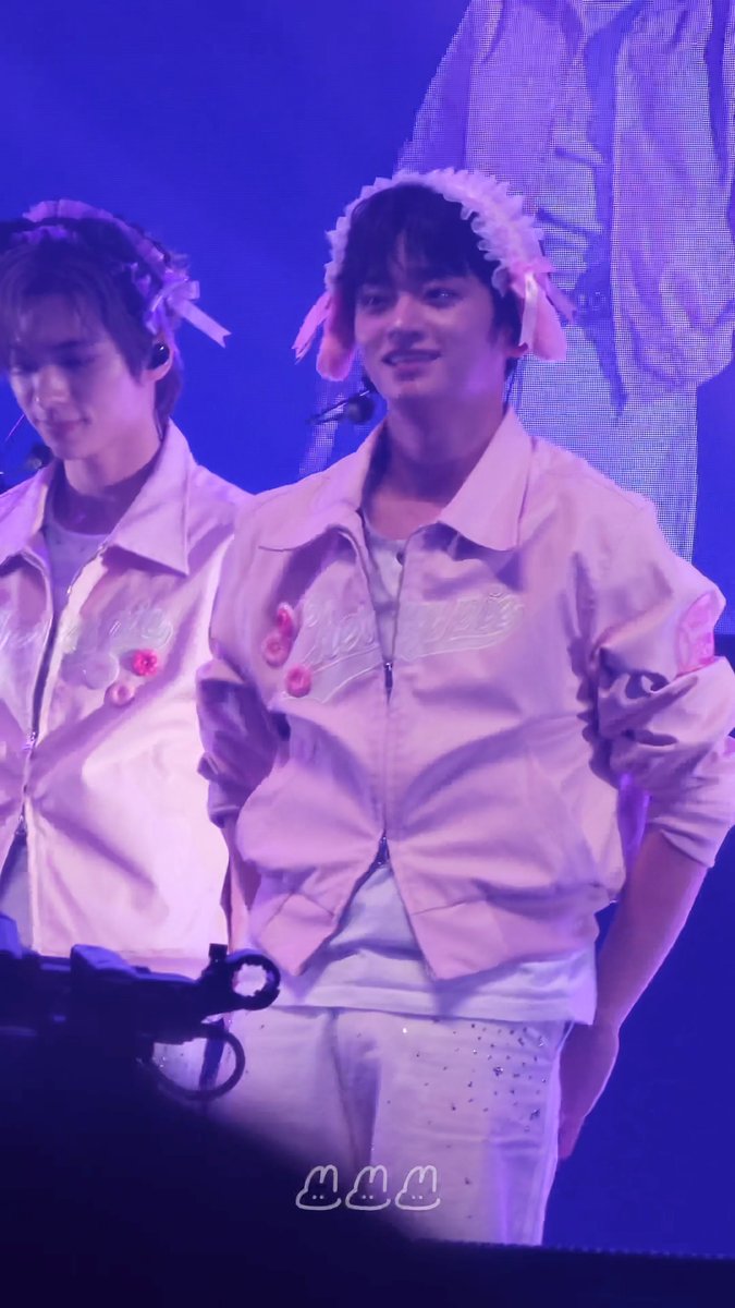 Two male K-pop performers stand on stage in matching pink satin jackets with bunny ear headbands, one smiling at the camera under purple lighting.