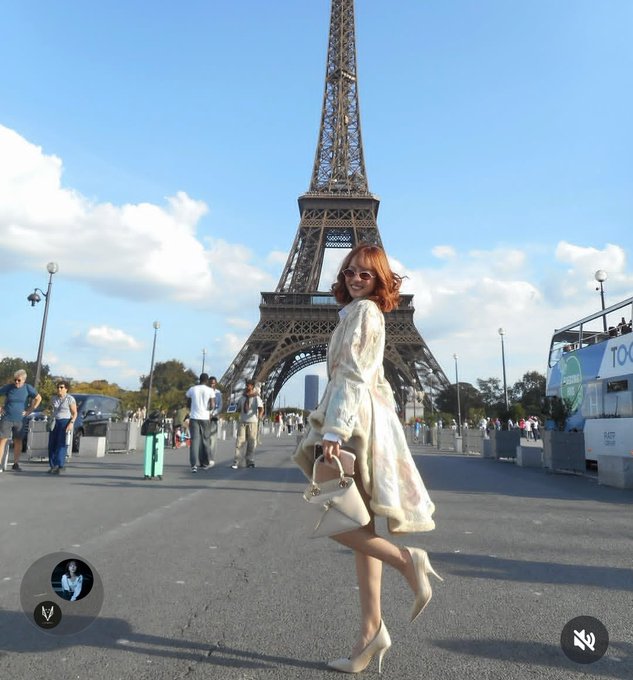 A woman with auburn hair wearing a floral dress, sunglasses, and beige heels posing in front of the Eiffel Tower in Paris, appearing to be a screenshot from a social media filter.