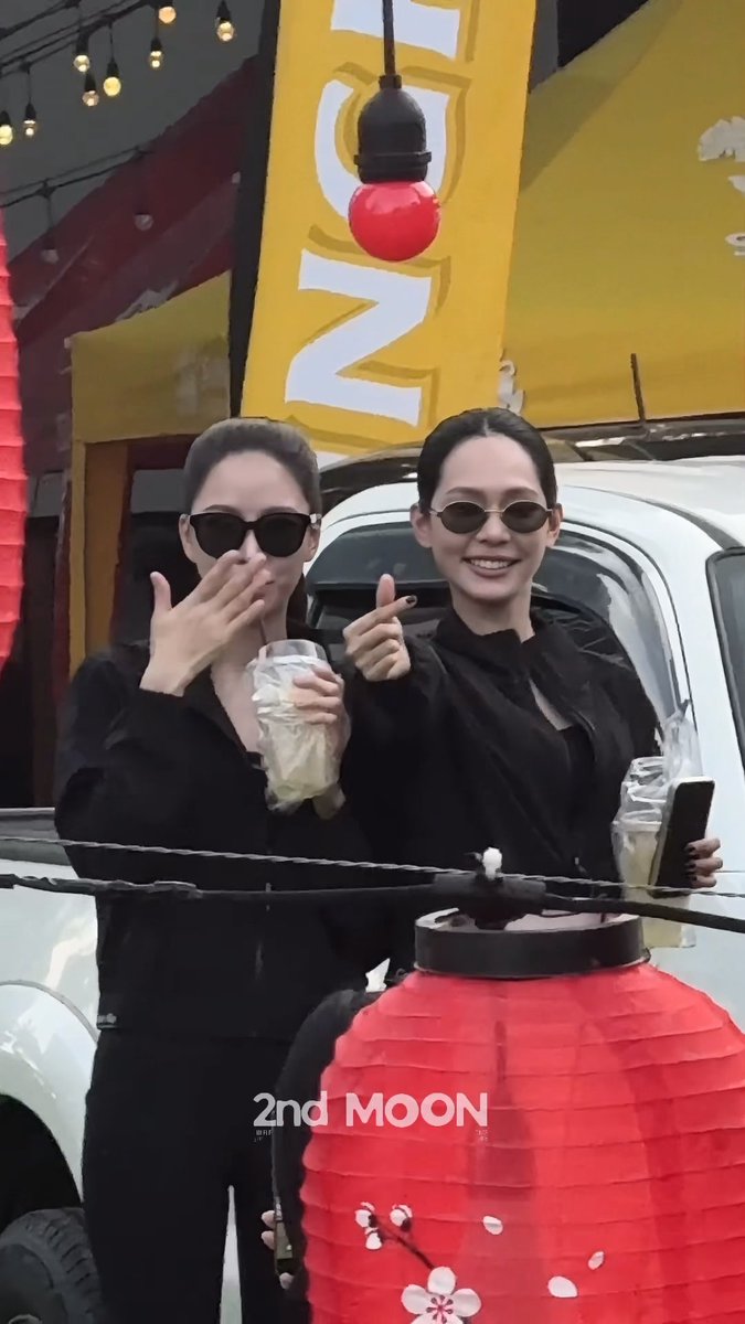 Two women in black outfits and sunglasses smile while holding drinks and posing behind a red lantern, with a yellow '2nd MOON' sign visible in the background.