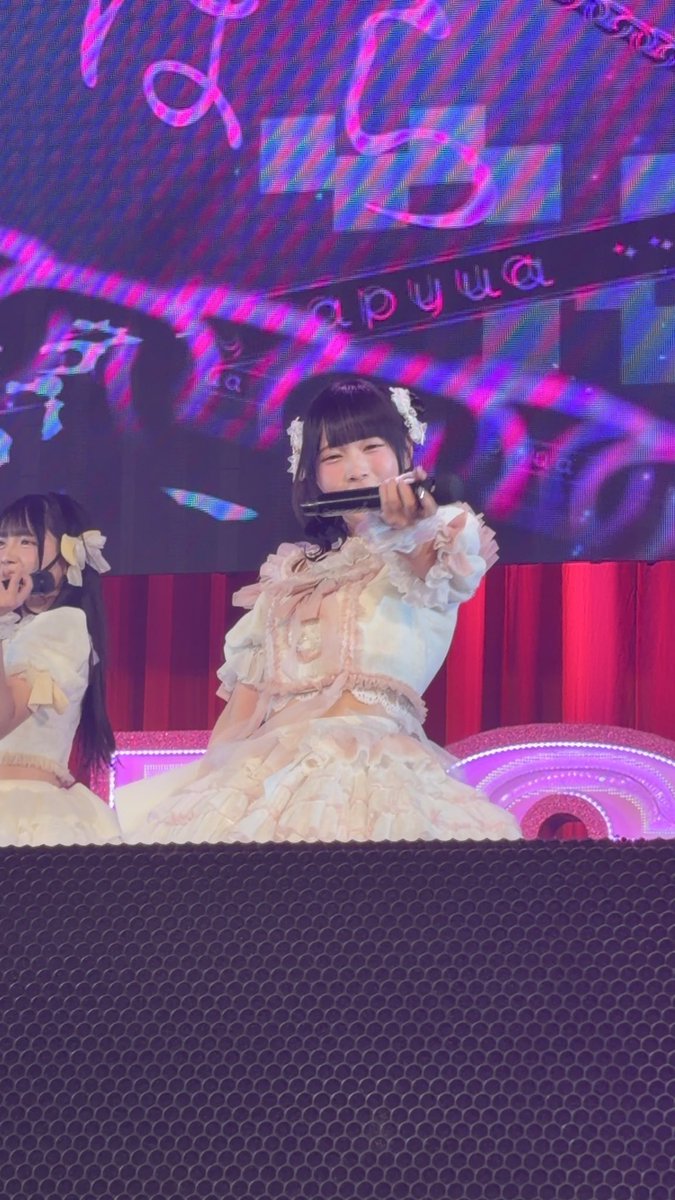 A young woman in a pink and white frilly idol costume singing into a microphone on stage, with colorful neon signage reading 'apjua' visible on the screen behind her.