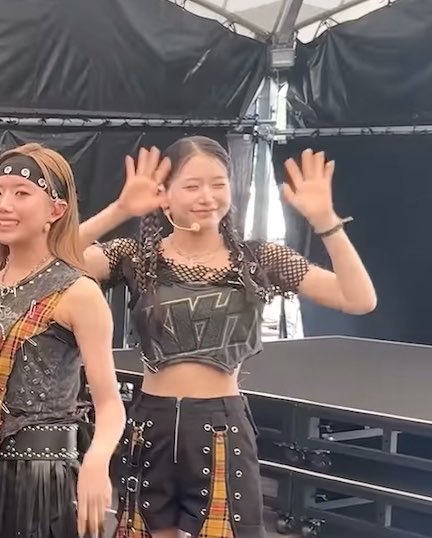 Two female performers wave to the camera on stage; one wears a black mesh crop top and patterned shorts, while the other wears a studded headband and a similar outfit.