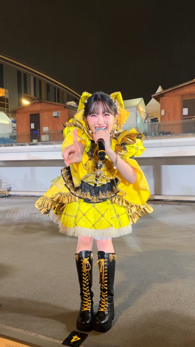 A smiling young woman performs on stage while holding a microphone, wearing an elaborate yellow and gold frilly dress with matching yellow-laced knee-high black boots.