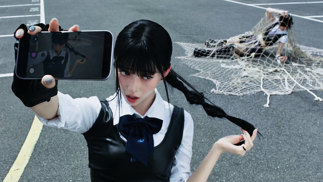 A young woman in a black and white school uniform holds the device up to take a selfie, while another figure in a similar outfit is caught in a white rope net on an athletic track.