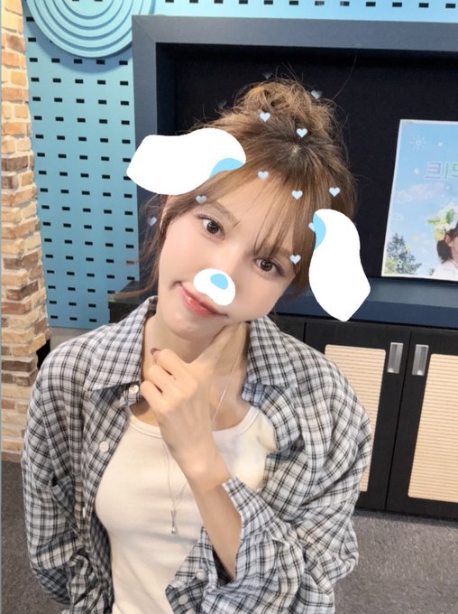 A young woman posing indoors wearing a plaid shirt over a white top, with cute animated cloud and heart face filters applied to her photo.