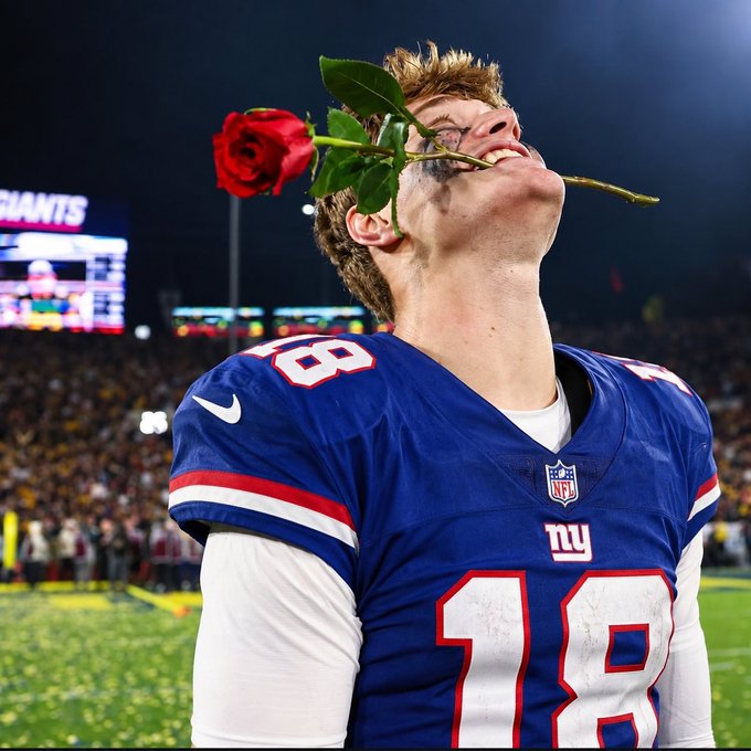 A New York Giants football player, wearing a blue jersey with number 18, bites down on a red rose in celebration on the field with confetti and a stadium scoreboard in the background.