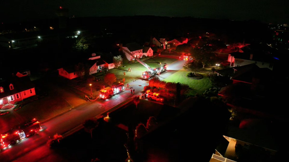 An aerial nighttime view of a residential neighborhood with multiple fire trucks and emergency vehicles responding to an incident, illuminated by red emergency lights.