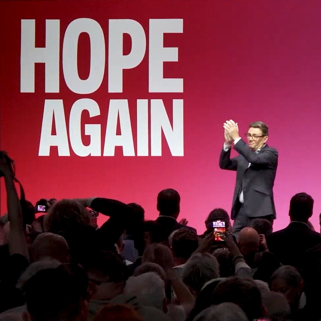 A man in a dark suit and glasses stands on stage applauding, with a large pink backdrop displaying the white text "HOPE AGAIN" behind him, as a crowd in the foreground holds up phones to capture the moment—a stark contrast to the post's demand for someone "peddling reality."
