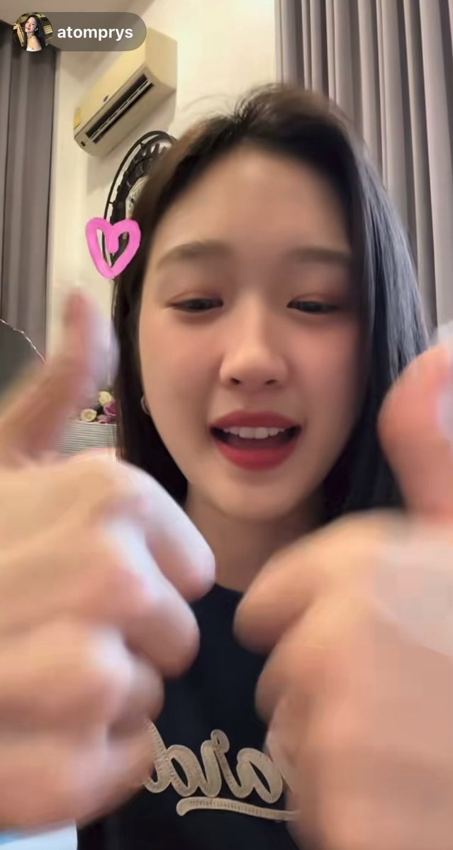 A young woman with long dark hair and red lipstick appears in a video call or live stream from user 'atomprys', smiling and reaching toward the camera with a pink heart filter.