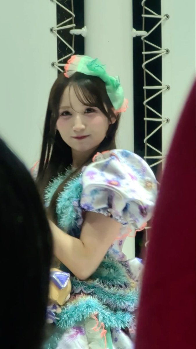 A young woman wearing a colorful pastel idol costume with puffy sleeves and a large green and pink hair bow smiles at the camera in what appears to be a backstage setting.
