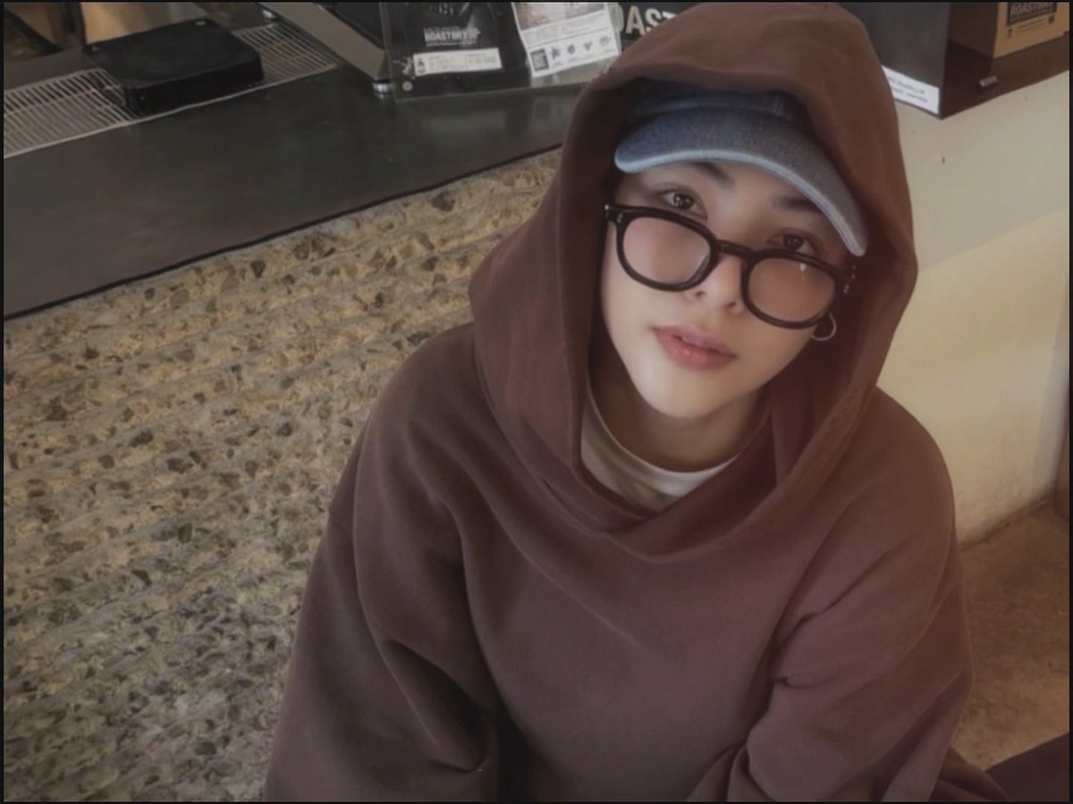A young person wearing a brown hoodie pulled up over a gray baseball cap and thick black glasses poses for a selfie in front of a stone-textured bar with bottles (including Jameson and Asahi) on the shelf behind them. The casual, confident "BOOM" vibe of the post matches their relaxed, slightly smirking expression and trendy layered streetwear look.