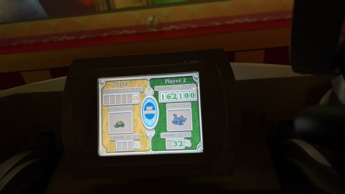 A photo taken at an arcade showing a competitive two-player game result screen, with Player 2 winning by a large margin (162,100) and Player 1 trailing with 32% scoring accuracy. The Japanese caption expresses the player's frustration with the lopsided loss and their wish to improve and play against real opponents.