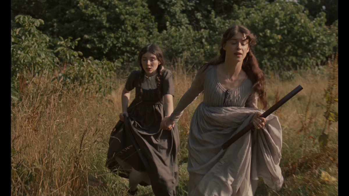 Two women in period dresses—one in a flowing white gown carrying a rifle, the other in a brown dress—run urgently through tall grass toward the camera, evoking a tense moment from the film adaptation of Jane Austen's *Sense and Sensibility*.