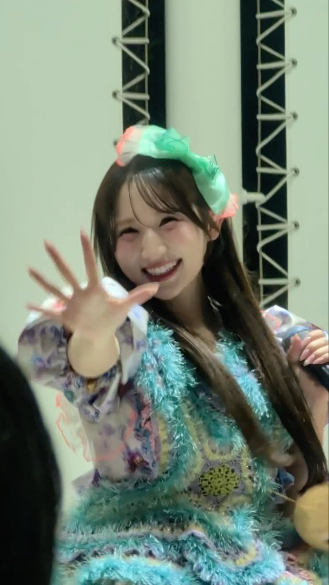 A smiling young woman with long dark hair and a colorful green bow headband waves at the camera, wearing a patterned outfit with turquoise tinsel trim.
