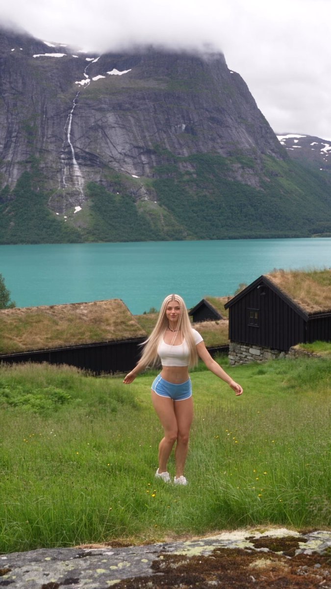 A blonde woman in a white crop top and blue shorts standing on grass in front of a stunning turquoise lake and dramatic mountain landscape.