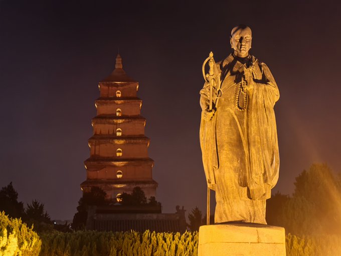 A nighttime photograph featuring an illuminated bronze statue of a monk/figure in robes in the foreground, with a lit multi-tiered pagoda in the background.