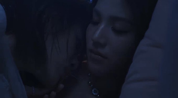 Close-up cinematic shot of two people in an intimate embrace in a dimly lit, blue-toned setting.
