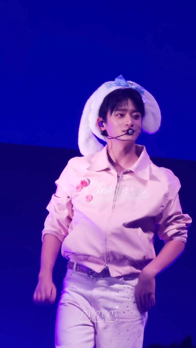 A young male performer on stage wearing a pink zip-up jacket, white pants, and a fluffy white hat with bunny ears, illuminated by blue stage lighting.