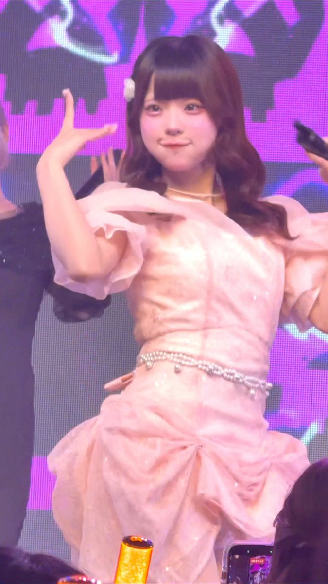 A young performer with long brown hair and bangs smiles on stage, posing in a frilly pink dress with a jeweled belt against a vibrant purple backdrop. The caption "巻き下ろしのふわふわガチ好き" ("I seriously love the fluffy, down-rolled hair") highlights the voluminous, wavy curls of her hairstyle, which match the soft, ruffled texture of her dress.