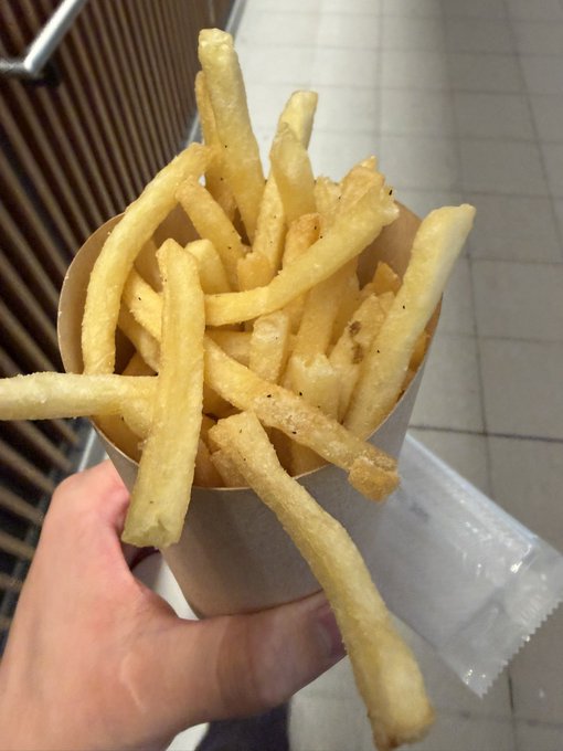 A hand holding a paper cone filled with golden, seasoned french fries in what appears to be a fast food setting.