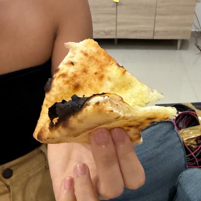 A person holds a folded slice of pizza in a relaxed, casual grip between their fingers. The post playfully notes how the girl holds the pizza, suggesting a unique or unconventional way of eating it.