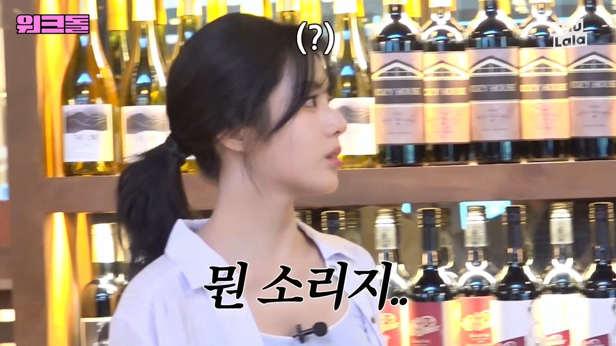 A woman with a confused expression stands in front of a wine rack, with Korean subtitles reading "뭔 소리.." ("What are you talking about..") and a "(?)" above her head. The image captures her flustered reaction to an awkward or unexpected question, matching the post's humorous depiction of a conversation gone sideways.