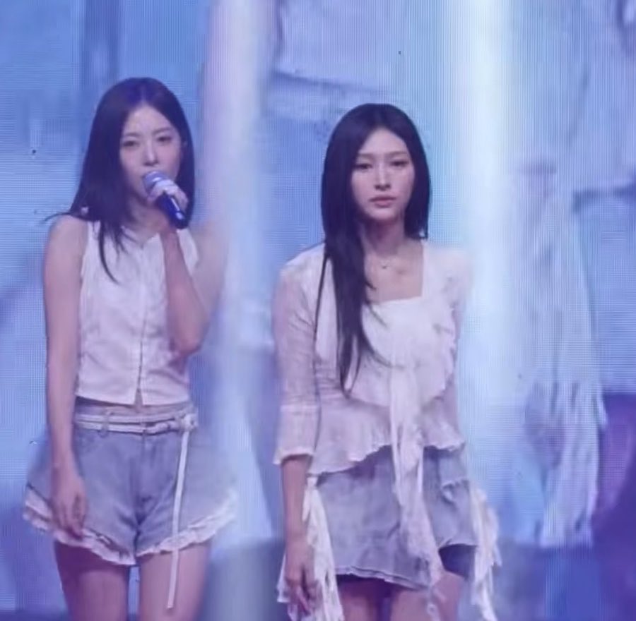 Two female K-pop performers on stage in coordinated white tops and gray skirts, with one singing into a microphone.