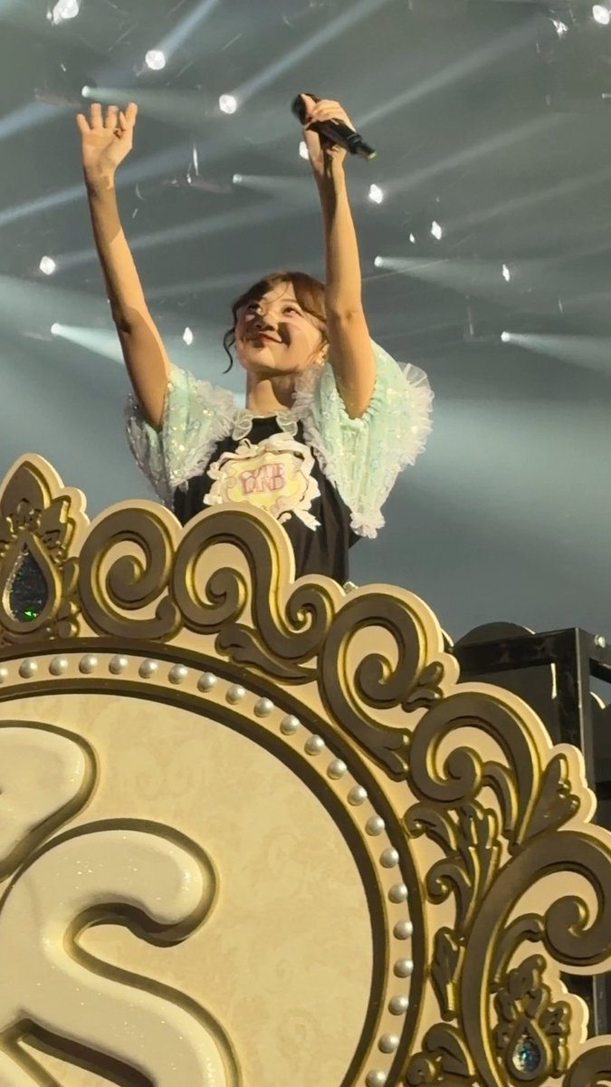 A performer, identified by the post as Itakura Kana from the idol group 26時のマスカレイド (whose shirt reads "26M"), triumphantly raises her arms with a microphone while standing atop an ornate gold-decorated stage prop during a concert moment.