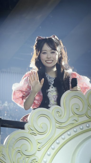 A smiling young woman with long dark hair, wearing a pink floral dress and a cat-ear headband, waves warmly while holding a microphone on an ornate stage with decorative yellow scrollwork, appearing to be at a concert or idol event, matching the caption celebrating "Manaka Sano" (佐野愛花).