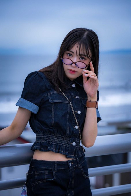 A portrait of a young woman wearing purple-tinted oval sunglasses and a dark denim crop top, standing by a railing with the ocean in the background.