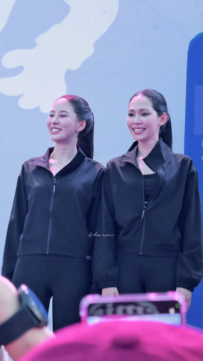 A photograph of two smiling women with slicked-back ponytails wearing matching black zip-up jackets, standing in front of a blue and white backdrop, possibly at a fan meeting or event, with a watermark reading 'thaaiin'.