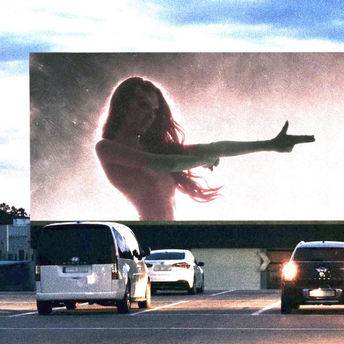 A billboard advertising for what appears to be a perfume or fragrance features a silhouette of a woman with long flowing hair, her arms extended outward, set against a glowing pinkish-purple background. Three cars, including a minivan and a sedan, drive past on the road below under a partly cloudy sky, while the figure's featureless face could be mistaken for wearing a mask.