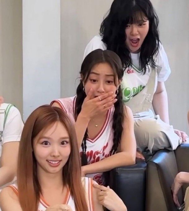 Three young women are captured in a candid moment, with one covering her mouth in surprise and another behind her with an open-mouthed excited expression.