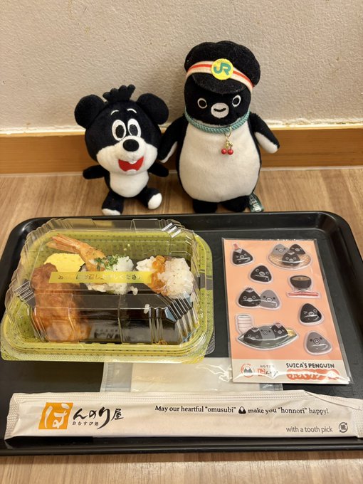 A Japanese bento meal features a clear plastic container of onigiri and other food, accompanied by two plush toys (a black bear and a penguin with a JR hat), stickers, and chopsticks from 'Honnori' stating 'May our heartfelt "omusubi" make you "honnori" happy!'