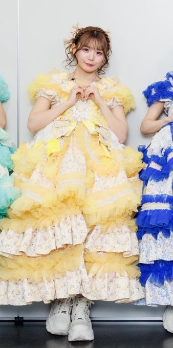 A young woman poses in an elaborate yellow frilly idol dress with floral patterns, white boots, and a matching hair accessory, forming a heart shape with her hands.