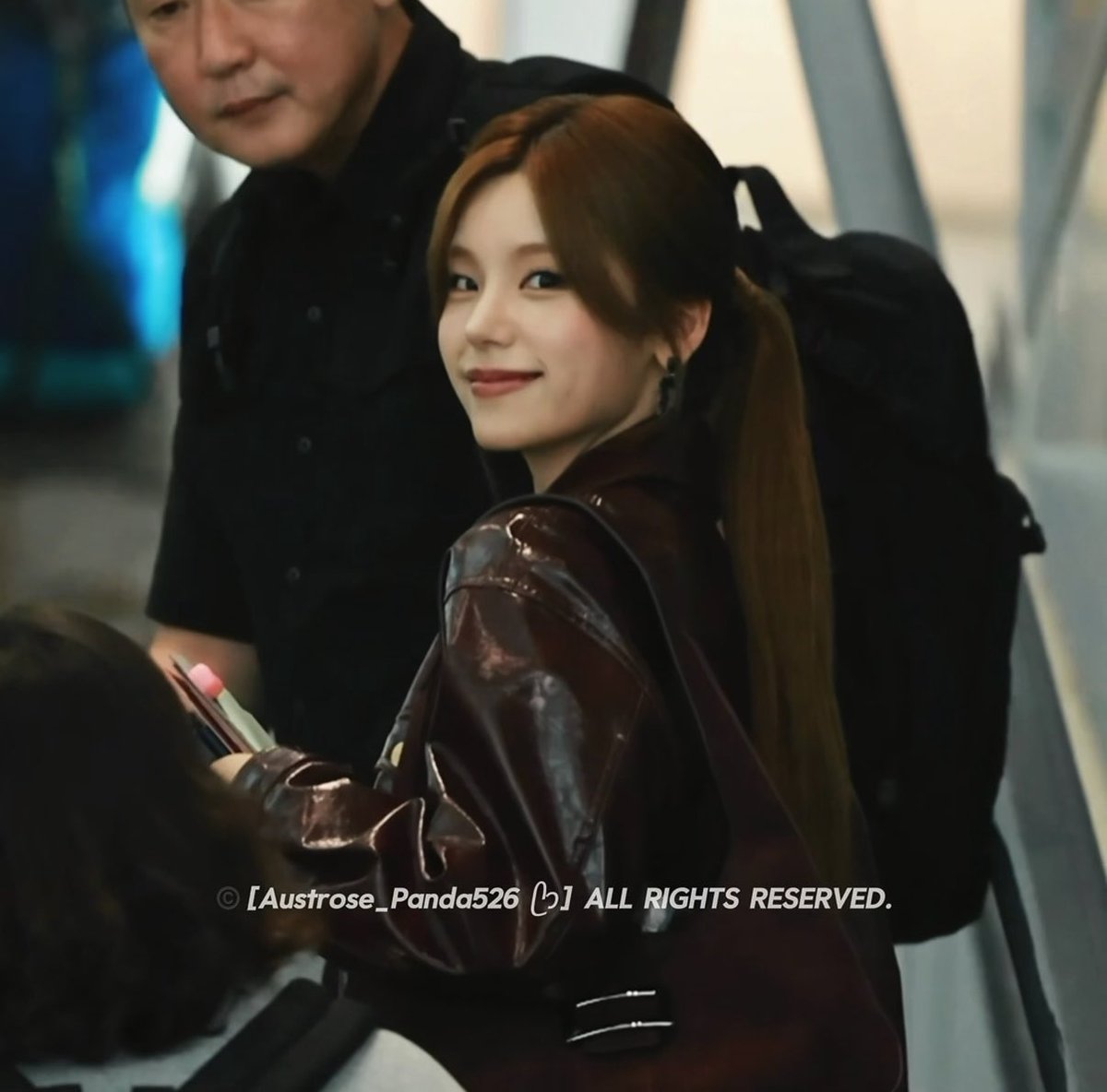 A young woman with long brown hair in a ponytail wearing a dark burgundy leather jacket, smiling as she looks over her shoulder, with a security guard visible behind her.