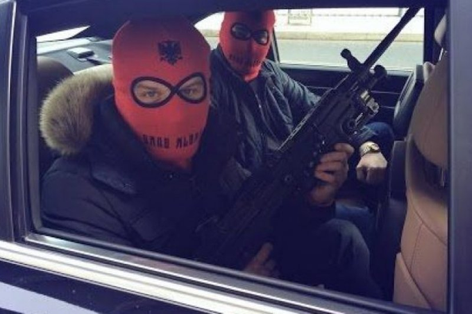 Two people wearing red ski masks sit inside a car, with the passenger holding a rifle.