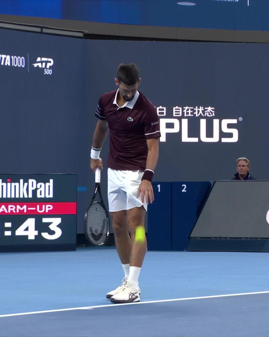 Djokovic is captured on court wearing a maroon Lacoste polo and white shorts while bouncing a ball during warm-up, with signage visible for "ATP 1000," "ThinkPad," and "PLUS" in the background. The image aligns with the post's praise of his stylish Lacoste outfit.