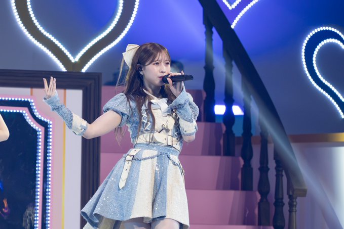 A young woman with long brown hair performs on stage in a sparkly blue and white outfit, singing into a microphone while raising her left hand, with decorative lighting and a staircase in the background.