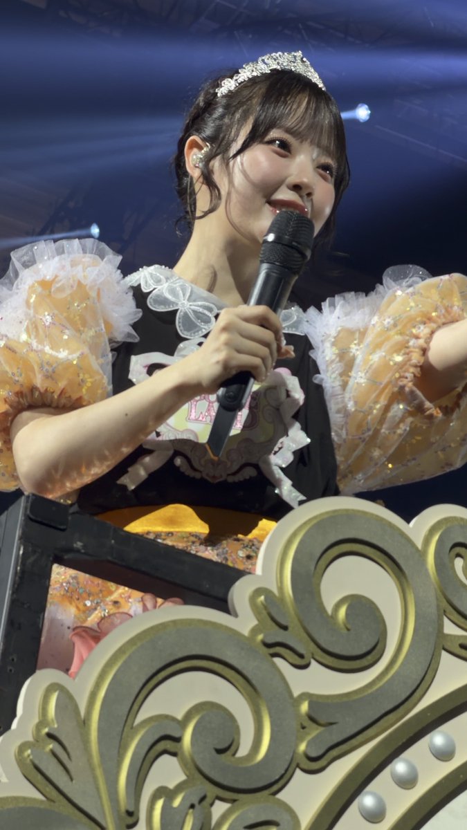 A smiling performer wearing a tiara and an elaborate black and yellow outfit with ruffled sleeves holds a microphone while seated behind an ornate gold decoration.