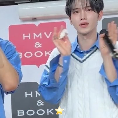 A young man in a blue shirt and white sweater vest clapping, with an 'HMV & BOOKS' logo visible in the background.