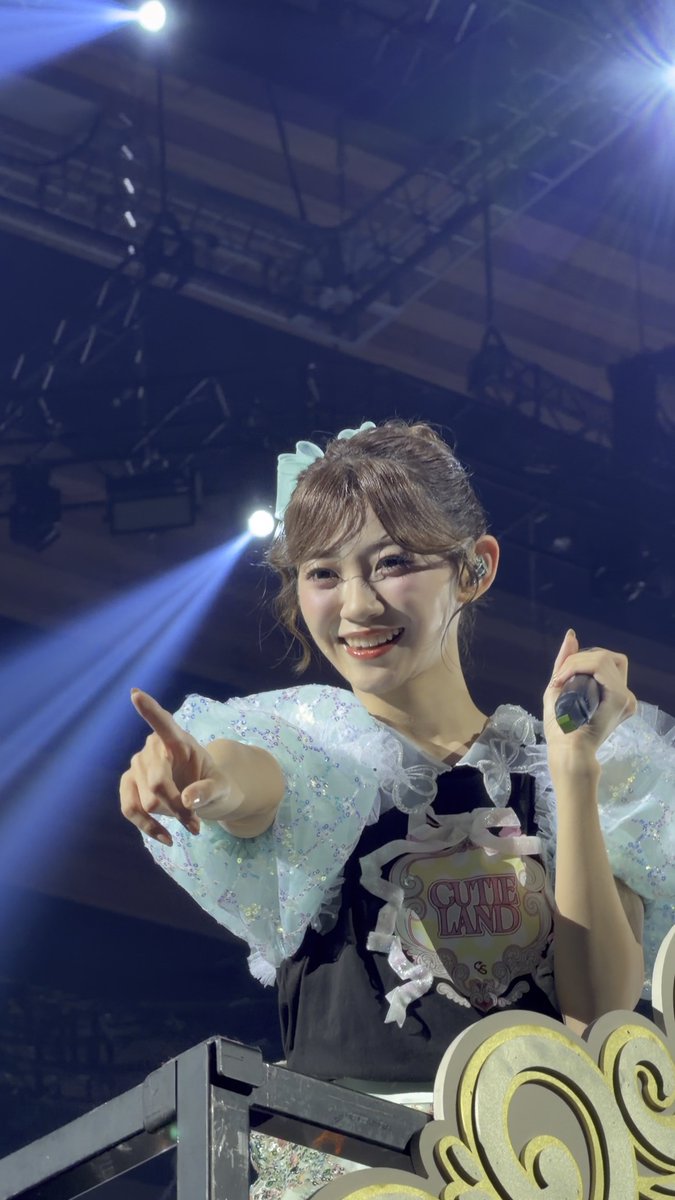 A young performer on stage smiling and pointing toward the camera while wearing a frilly black and pastel outfit with a 'CUTIE LAND' patch and holding a microphone.