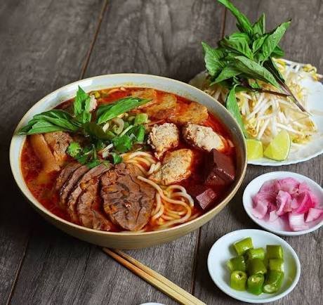 A bowl of Vietnamese-style beef noodle soup (pho) served with a side plate of bean sprouts, basil, lime, pickled onions, and sliced green chilies on a wooden table.