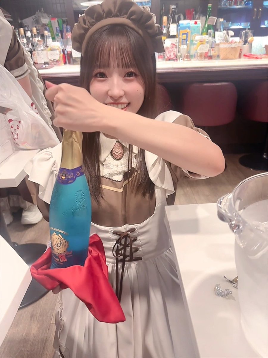 A smiling young woman wearing a brown and white maid outfit and headband holds up a blue bottle of Moon Blue champagne at what appears to be a maid café.