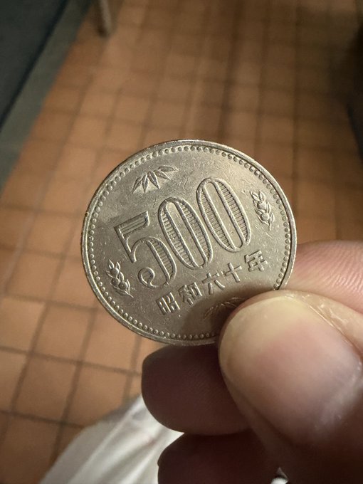 A close-up of a Japanese 500 yen coin featuring the inscription "500" and Japanese characters meaning "Heisei 16" (2004) being held between fingers. The post relates by criticizing the coin for being incompatible with modern vending machines, parking meters, and convenience store registers, suggesting it's become obsolete and should be removed from circulation.