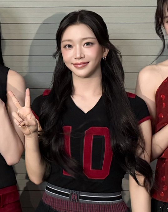 A young woman with long dark wavy hair poses making a peace sign, wearing a black shirt with red '10' lettering and a red plaid skirt.