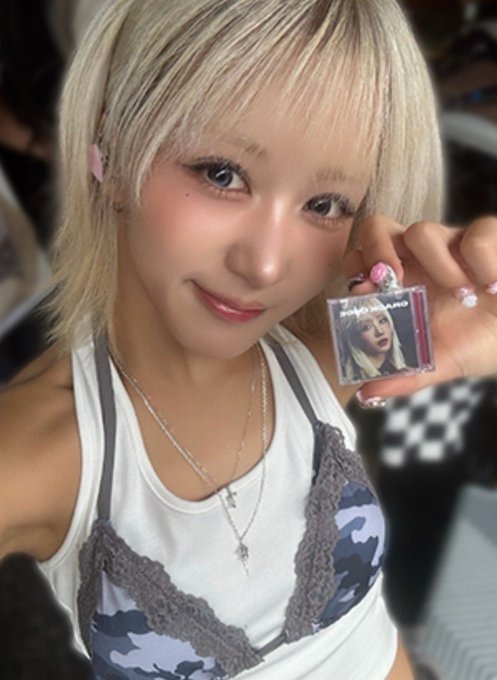 A blonde woman with a nose ring wearing a camo top smiles while displaying a small keychain or photo charm that reads 'May' with her own image on it.