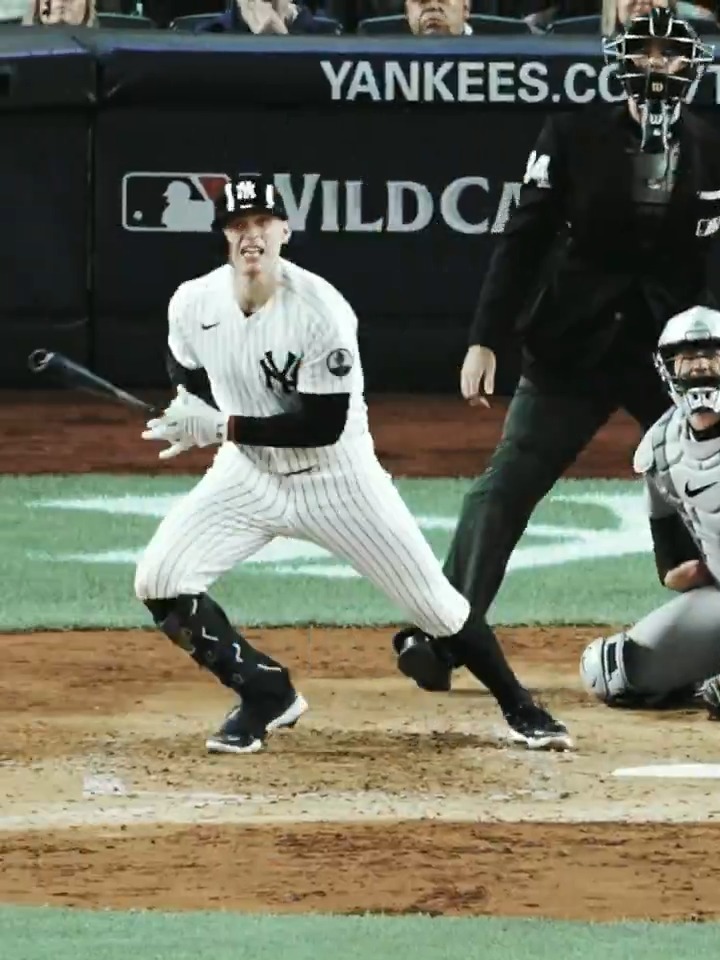 A New York Yankees baseball player in pinstripe uniform completes a swing during a game, with the umpire and catcher behind him and 'YANKEES.COM' and 'WILDCARD' visible on the wall.