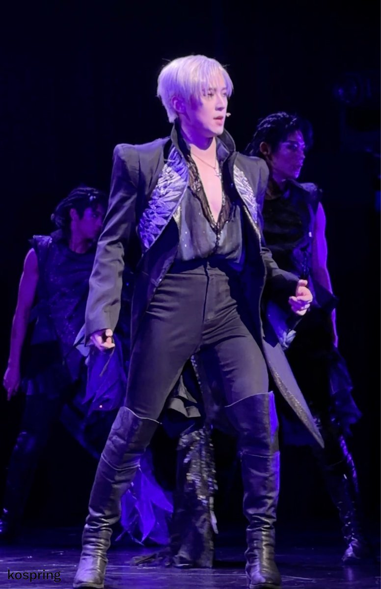 A performer with silver-purple hair stands center stage in a dramatic black outfit with ornate detailing, knee-high boots, and an open sheer shirt, holding a microphone under purple stage lighting while backup dancers pose in the background. The watermark "kospring" appears in the bottom left corner, and the post's hashtags (#고은성 #コウンソン) reference the performer Ko Eun-sung (コウンソン), suggesting this captures a memorable performance moment he or his fans particularly cherished.