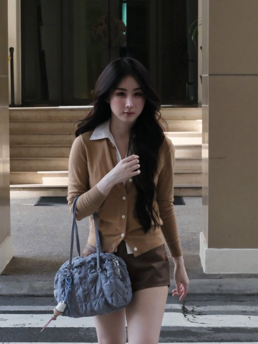 A stylish woman poses on a city street wearing a tan cardigan over a white collared shirt, paired with brown shorts and carrying a slouchy denim-blue handbag. The image captures a candid, fashion-forward moment, fitting for Yoko Apasra's Instagram post under the hashtag #YokoApasra.